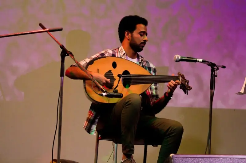 Oud player seated on stage between two microphones, playing under purple stage light