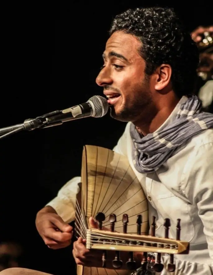 Oud player singing into a microphone while playing his oud on a dark stage