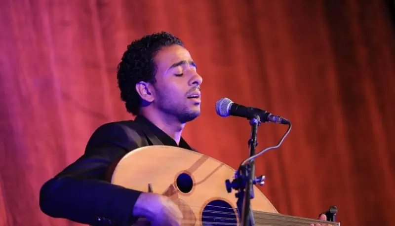 Oud player singing with closed eyes in front of a red stage curtain